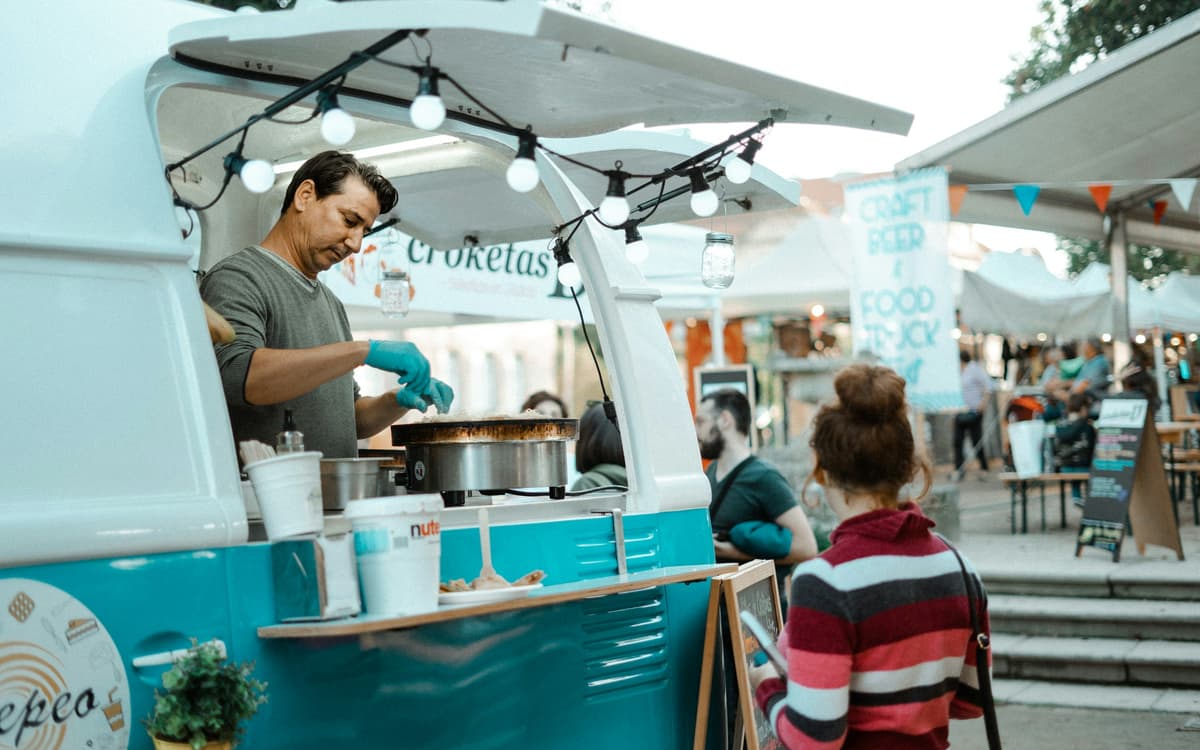 A cook works the window of a food truck while customers wait at an outdoor market.