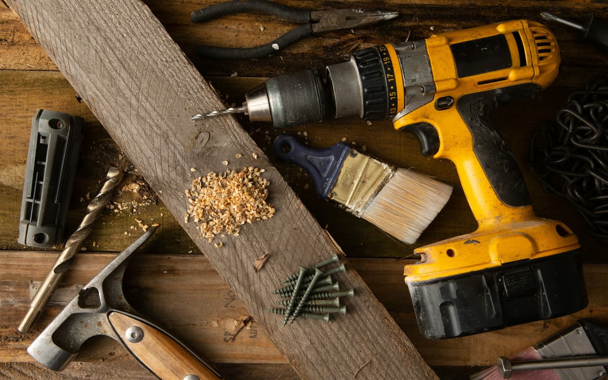 A cordless drill, hammer, paintbrush, screws and other hand tools on a wooden workbench.