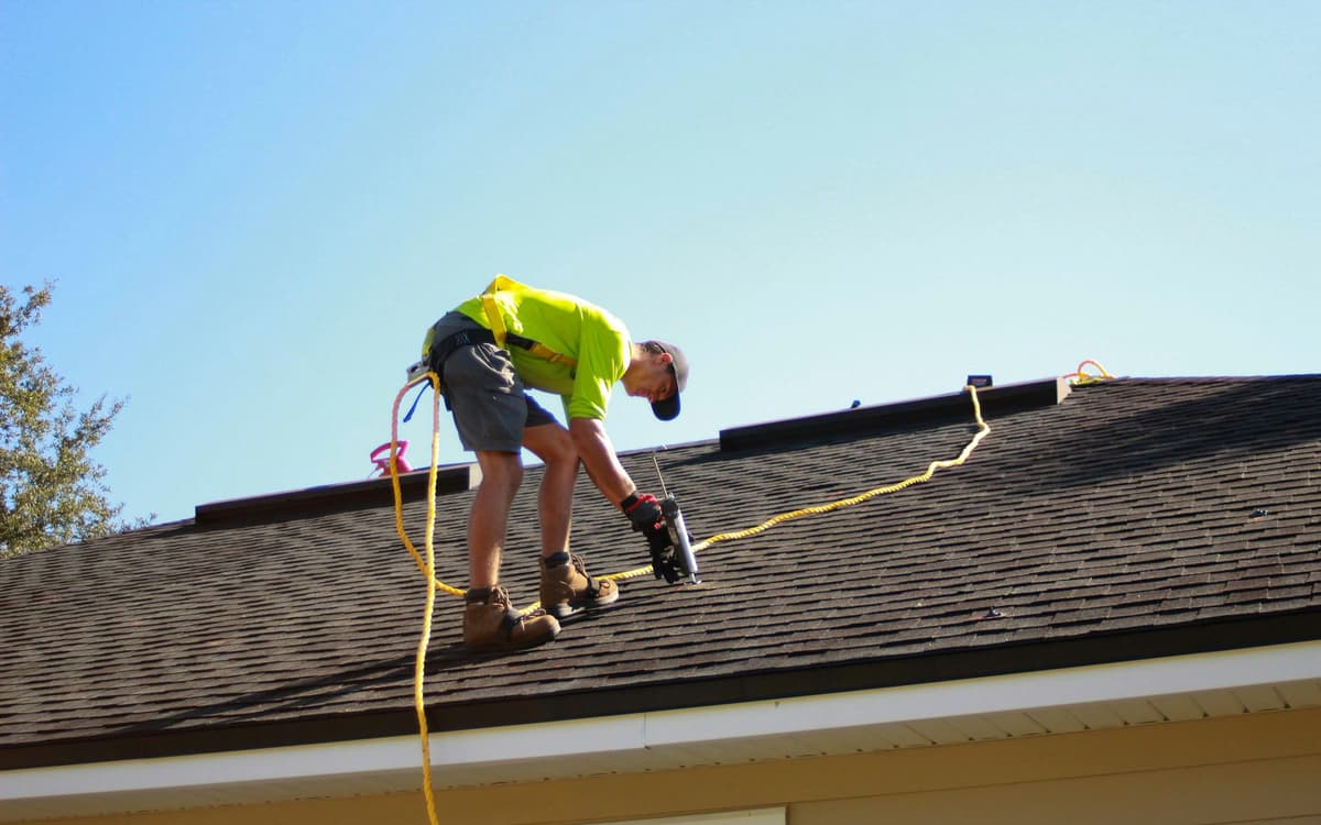 A roofer in a safety harness and rope fastens shingles on a sloped roof.